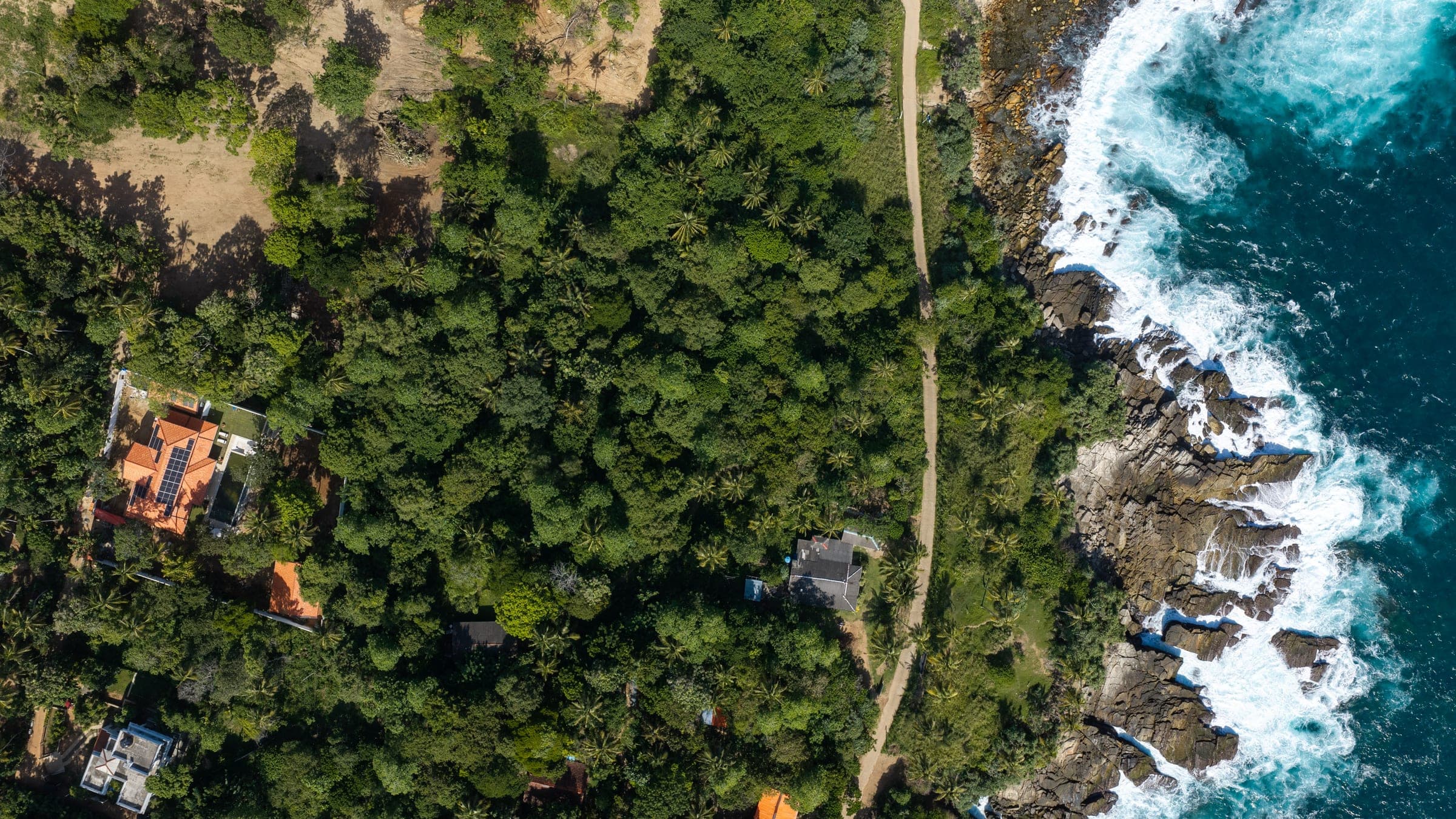 Aerial view of Kottamba Kanda's terracotta roofs and solar panels among the trees, with the surf of Sri Lanka's south coast alongside