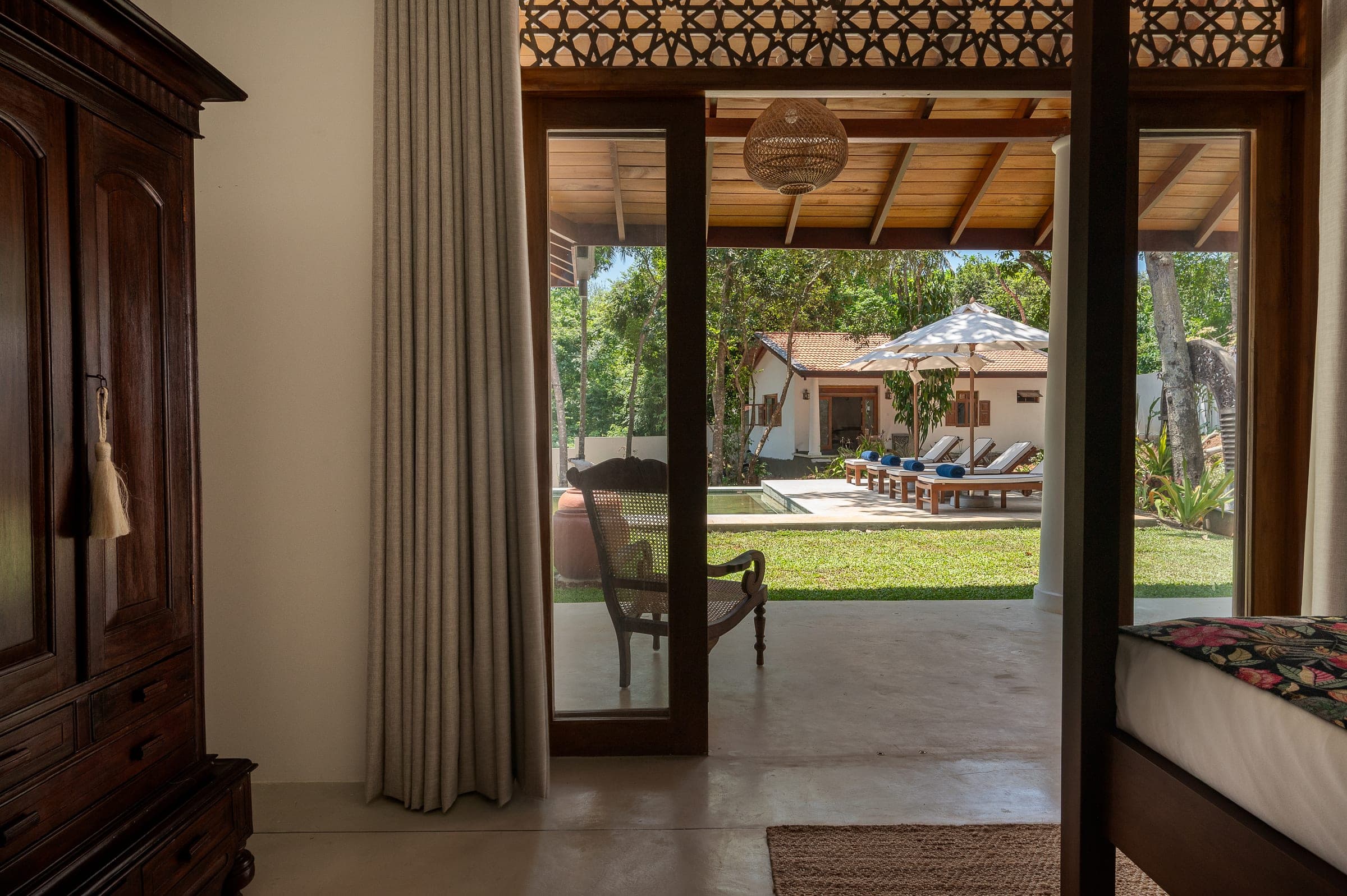 From Bedroom One through the open doors to a planter chair on the verandah, the pool and sun loungers beyond
